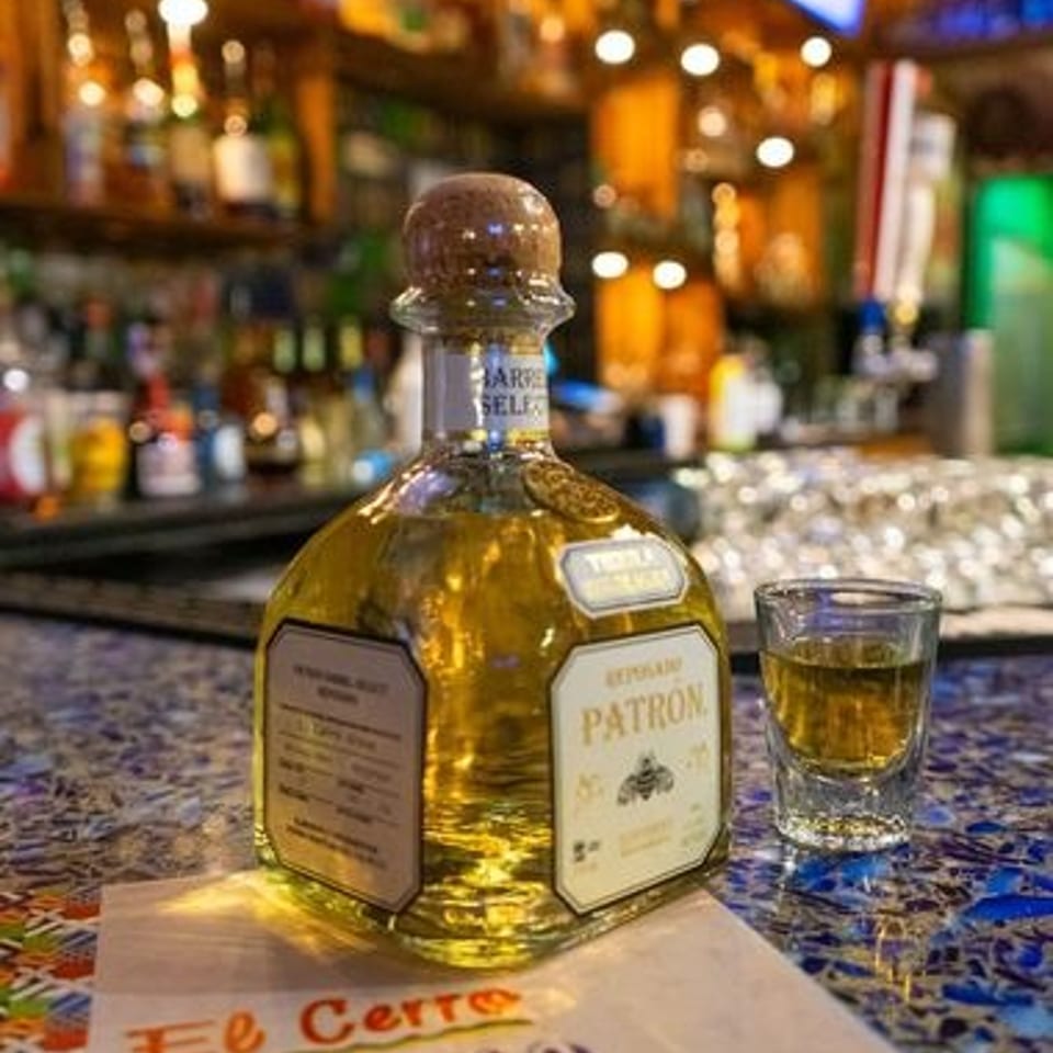 Patrón tequila bottle and filled shot glass on a colorful bar counter, with a warmly lit, blurred bar interior behind.
