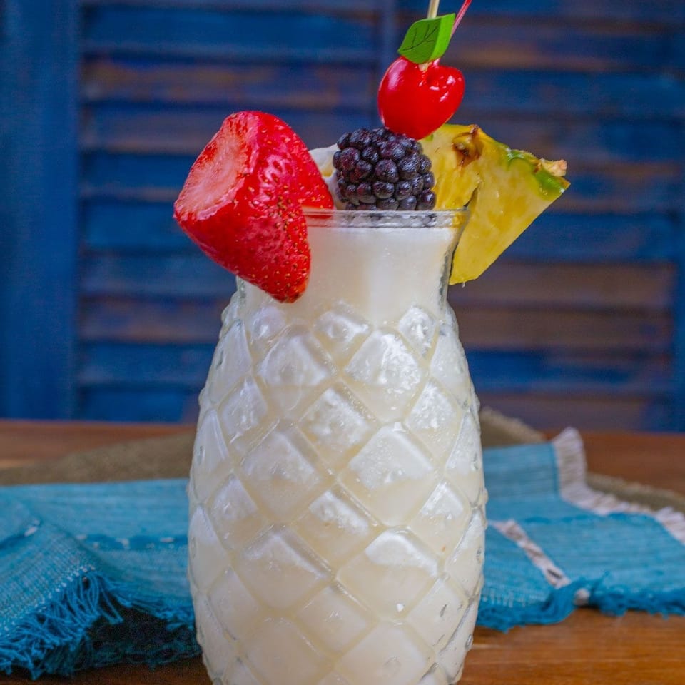 Creamy fruit cocktail served in a pineapple-shaped glass, garnished with strawberry, blackberry, pineapple, cherry, mint, and straws.