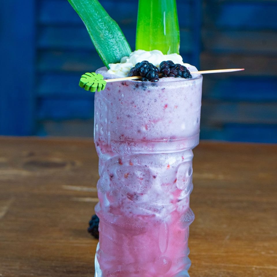 Layered pink berry cocktail topped with blackberries, whipped cream, green leaves, and a purple flower on a wooden table.