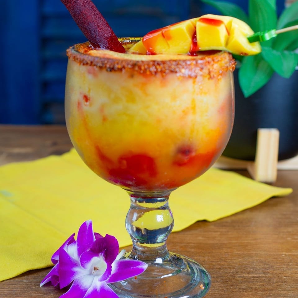 Tropical fruit cocktail in a chili-rimmed glass, garnished with mango, a tamarind straw, pineapple leaves, and purple orchids.