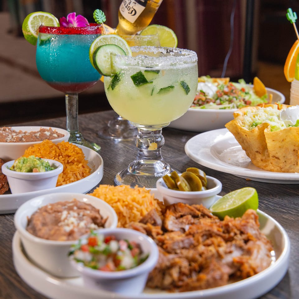 Mexican restaurant table with colorful margaritas, a beer bottle, rice, beans, guacamole, tacos, and garnished dishes.