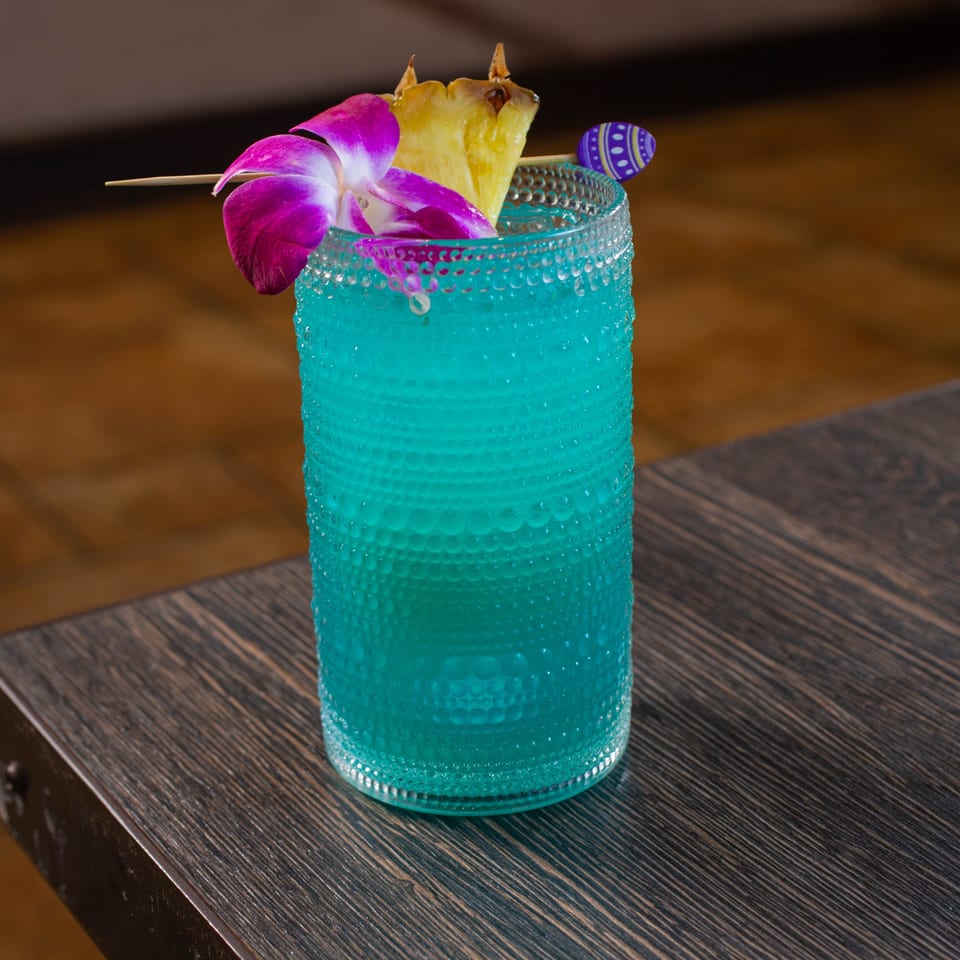 Bright turquoise cocktail in a textured glass, garnished with purple orchid, pineapple leaves, and a decorative pick on a restaurant table.