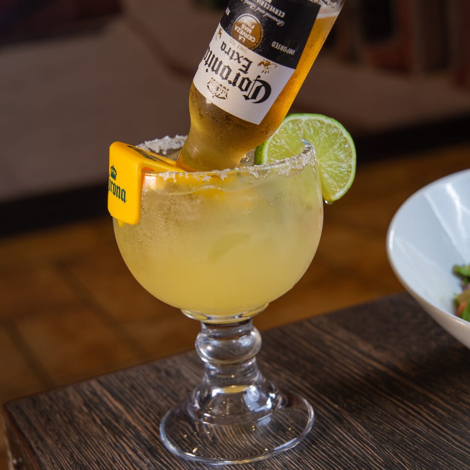 Salt-rimmed margarita with lime wedge and inverted Corona beer bottle, served in a stemmed glass on a wooden restaurant table.